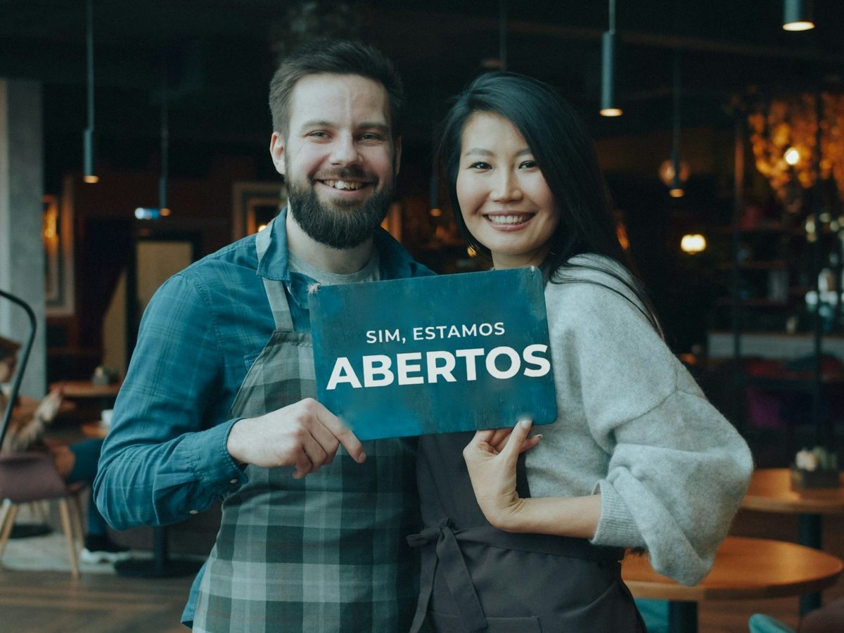 Dois sócios sorrindo na porta do café, segurando a placa de aberto